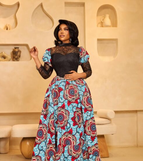 Elegant woman wearing a floor-length blue, red, and white African print gown with black lace bodice, posing in a beige showroom.