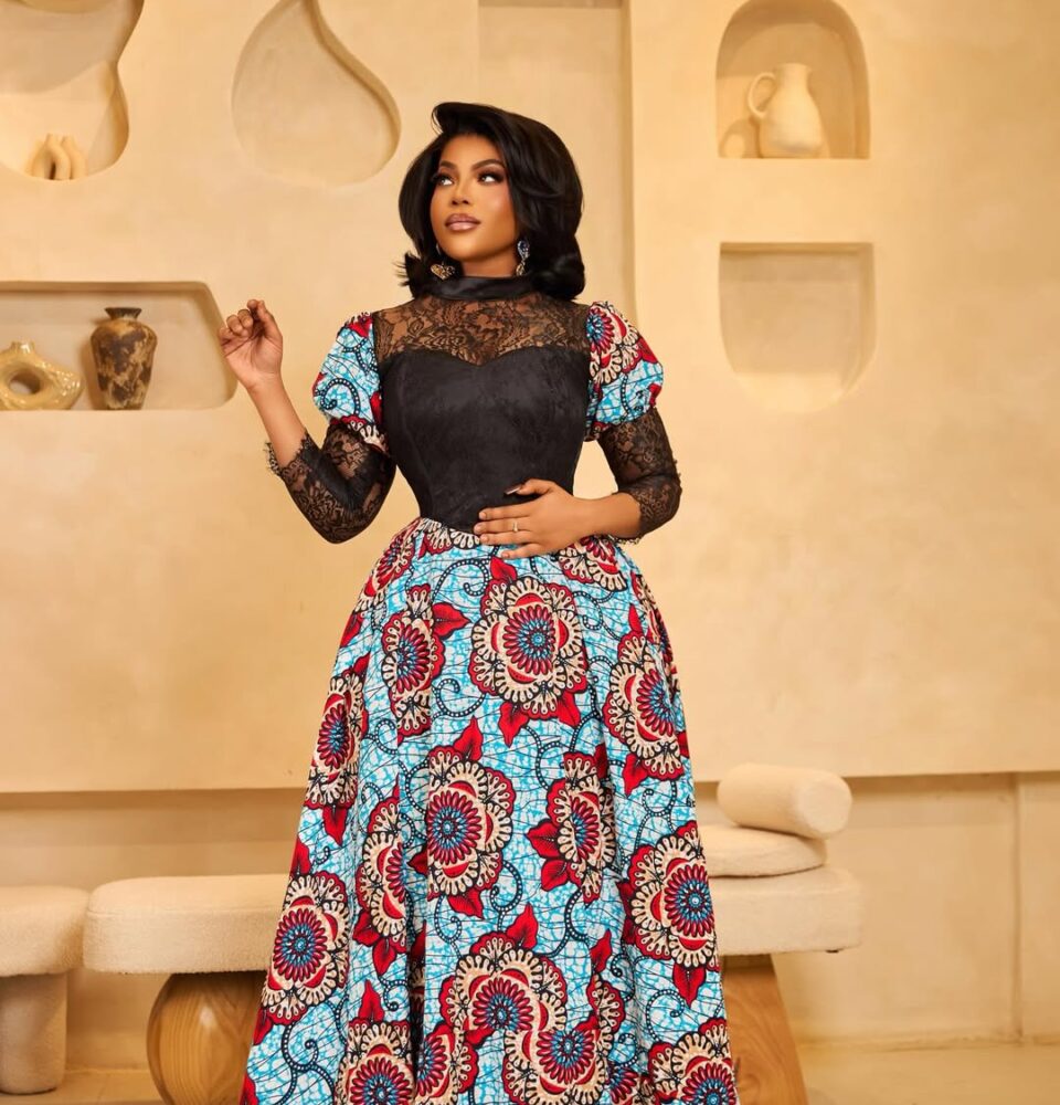 Elegant woman wearing a floor-length blue, red, and white African print gown with black lace bodice, posing in a beige showroom.